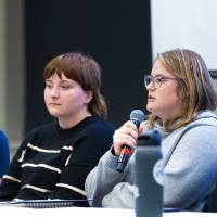 four college student panelists sitting at the front of the conference room, one female student speaking other others listen